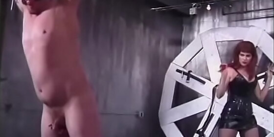 redhead chick in fishnet stockings ties the guy upside down to a big wheel and spanks him with a whip